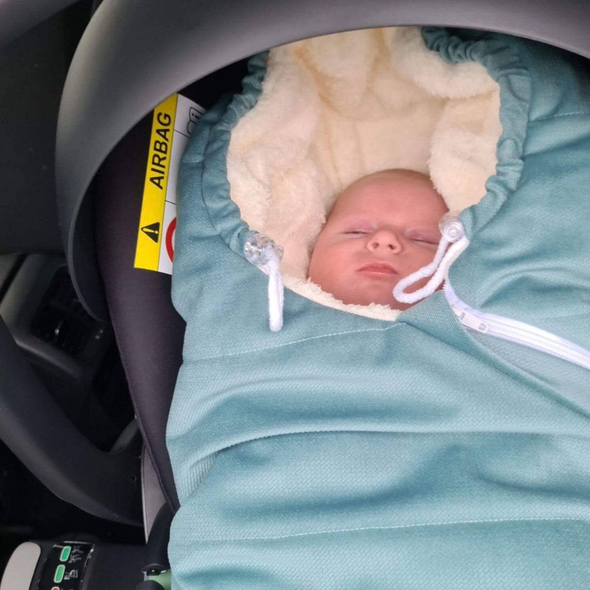 A sleeping newborn baby wrapped in a mint green baby footmuff with a cream fleece hood, resting inside a stroller pram, infant car seat.