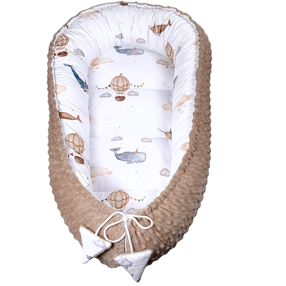 evcushy baby nest with a textured brown faux fur exterior and a white interior printed with watercolor-style hot air balloons, whales, and clouds, finished with two small white fabric bow accents at the bottom.