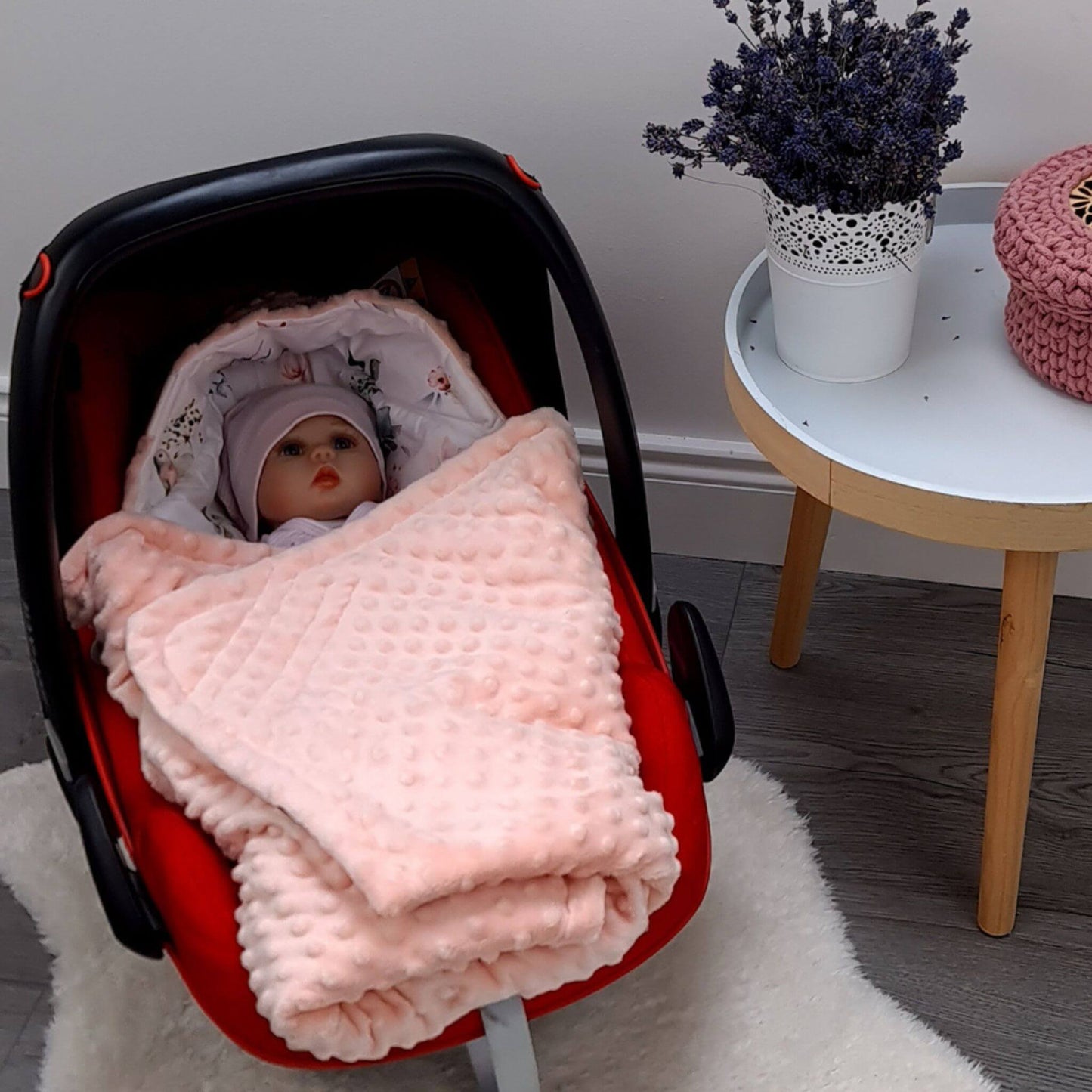 A baby blanket in a light pink color with a textured minky dot pattern, wrapped around a baby inside a red baby car seat, set on a white rug next to a small side table.