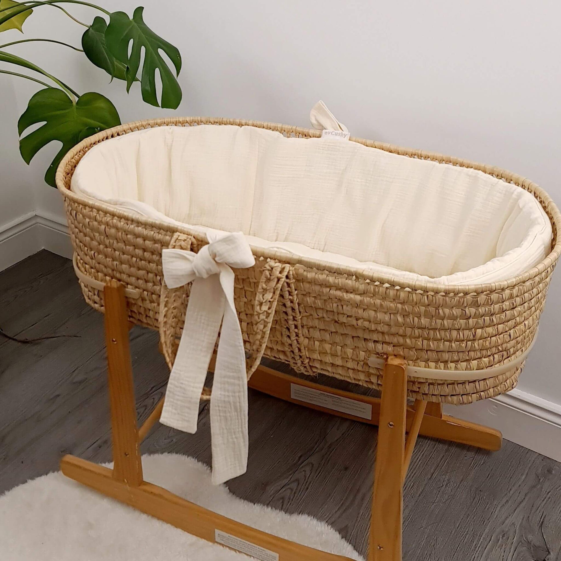 Wicker baby crib with cream liner mattress on a wooden stand in a room with a plant.