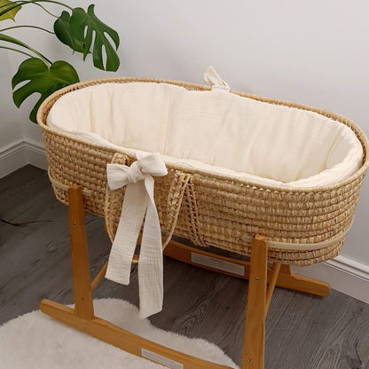 Wicker baby crib with cream liner mattress on a wooden stand in a room with a plant.