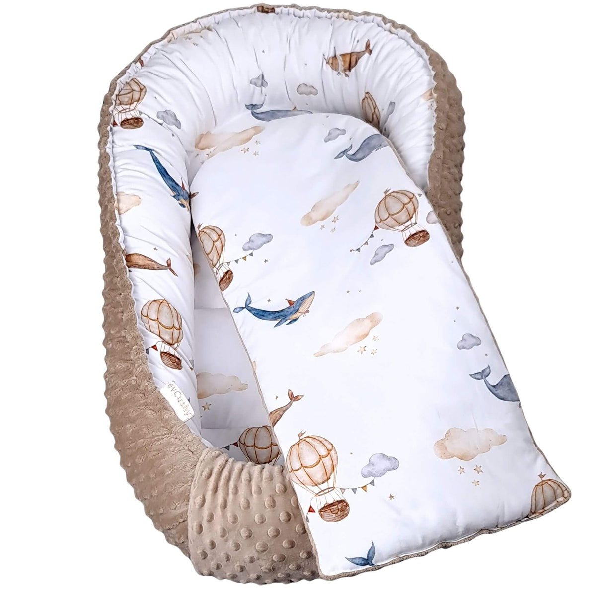 A baby bassinet or bassinet insert with a white printed fabric featuring blue whales, brown hot air balloons, and beige clouds, surrounded by a soft beige minky dot fabric border.