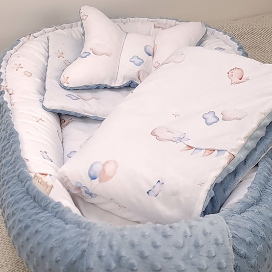 A baby bedding set arranged inside a soft padded bassinet or bed, featuring a white pillow and blanket printed with pastel clouds, balloons, and birds, with a light blue textured minky fleece border.