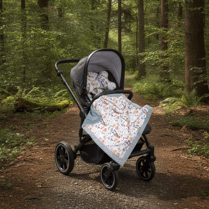 A stroller with a liner and head support pillow is parked on a forest path, with a light grey patterned blanket with animals design draped over the stroller seat.