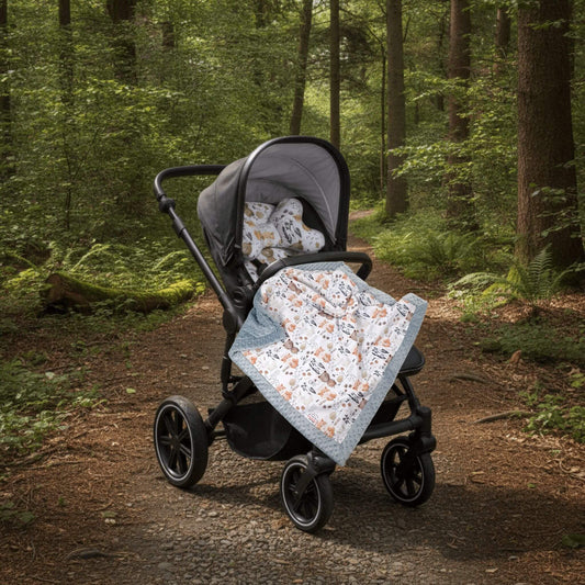 A stroller with a liner and head support pillow is parked on a forest path, with a light grey patterned blanket with animals design draped over the stroller seat.