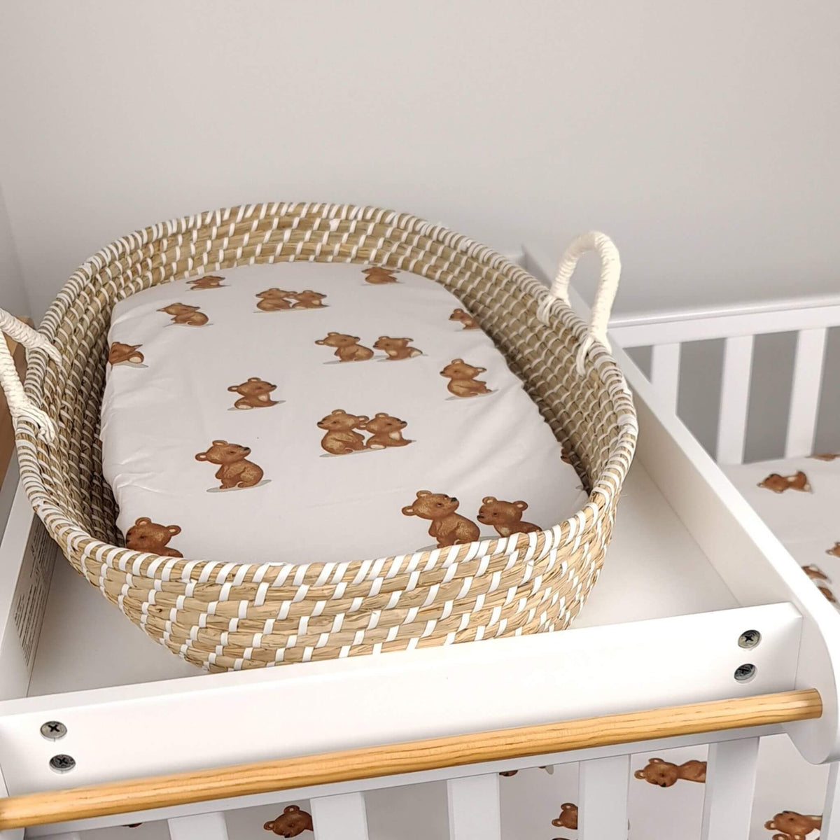 A woven natural fiber baby bassinet basket with white handles, holding a white fitted sheet printed with small brown teddy bear motifs, set inside a white baby crib.