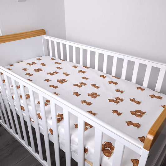 A white fitted crib sheet mattress with a printed pattern of small brown teddy bears, shown inside a white wooden baby crib with natural wood rails.