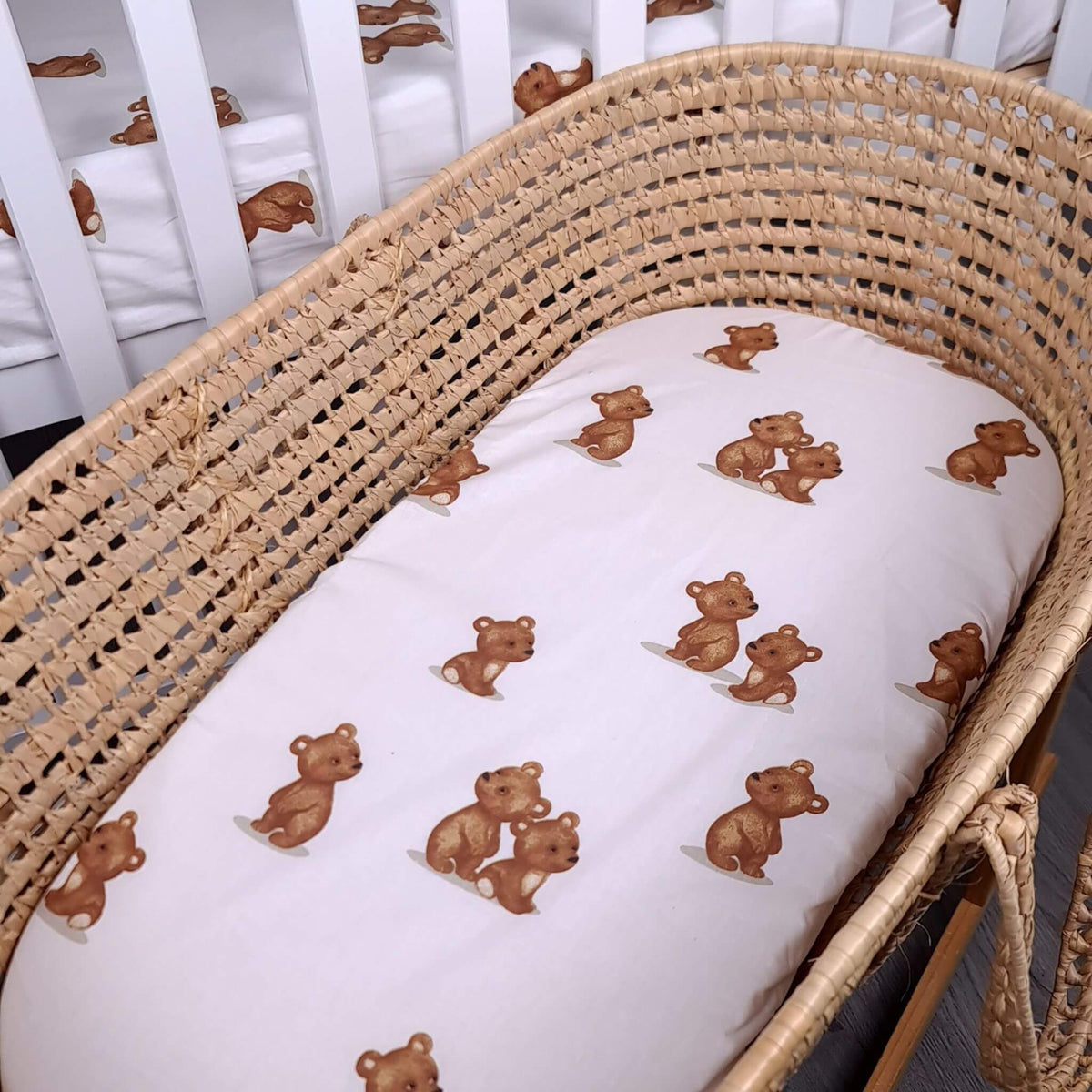 A woven rattan bassinet containing a white mattress with a repeating pattern of brown teddy bears, set against a white crib.
