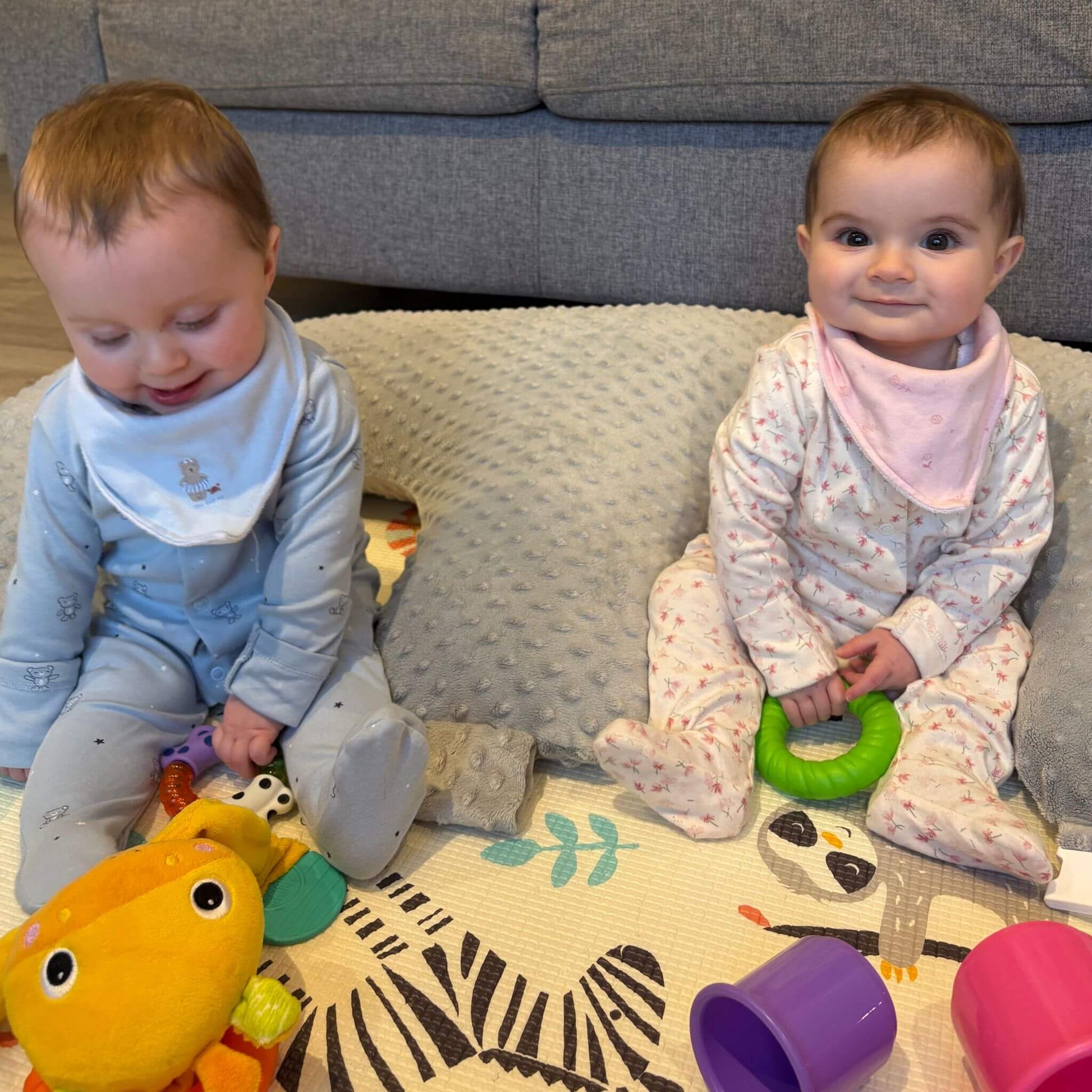 Two babies playing with toys supportrd by evcushy twin pillow in a living room.