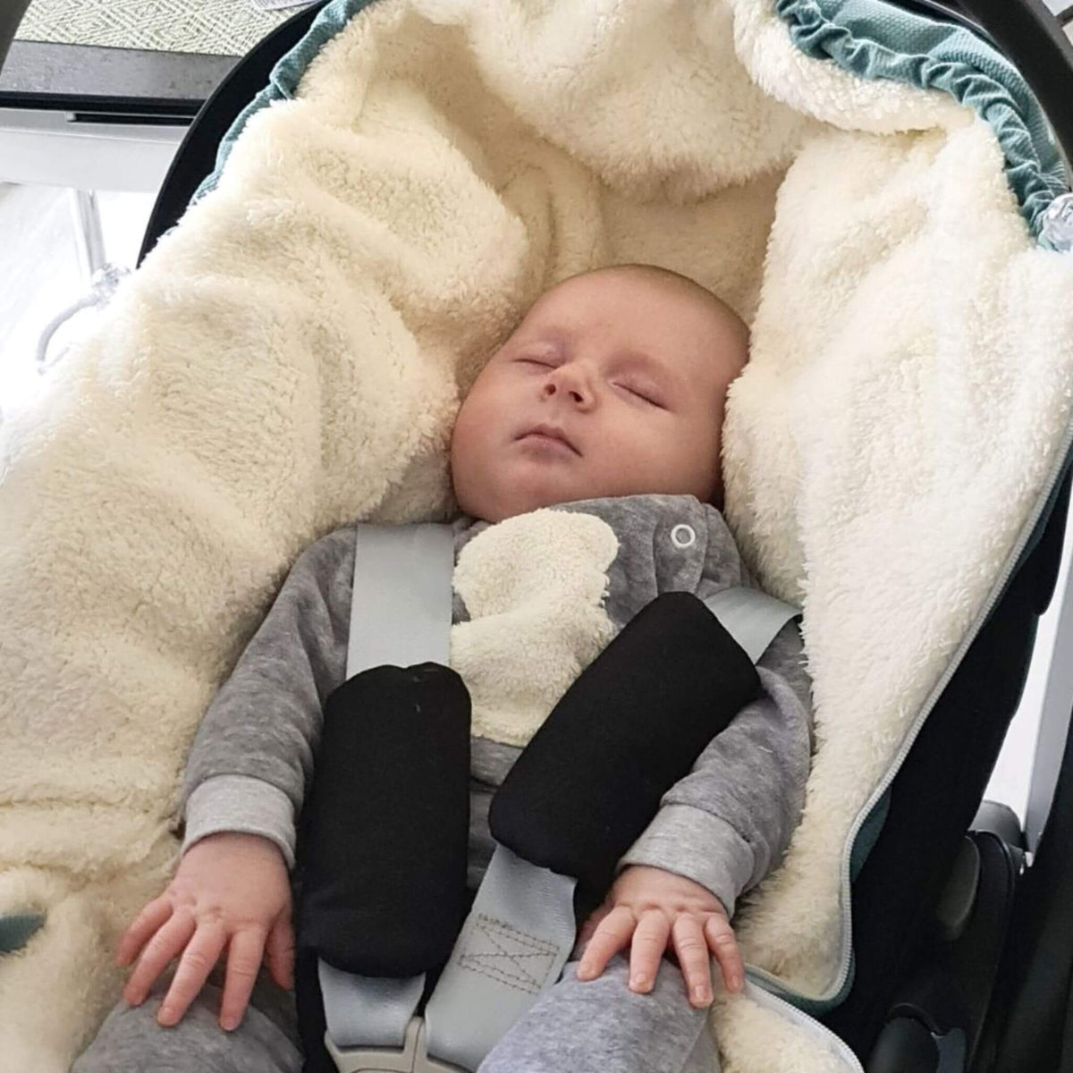 A sleeping baby wearing a gray outfit and lying in a black car seat, covered with a cream fleece footmuff
