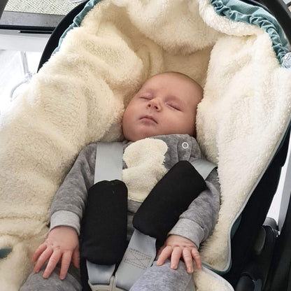 A sleeping baby wearing a gray outfit and lying in a black car seat, covered with a cream fleece footmuff